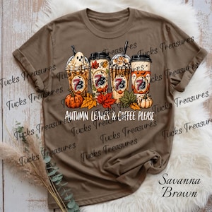 May include: A brown t-shirt featuring a graphic of four coffee cups decorated with autumn themes, including pumpkins, leaves, and a ghost. The text "AUTUMN LEAVES & COFFEE PLEASE" is printed below the cups. The shirt color is labeled as Savanna Brown.