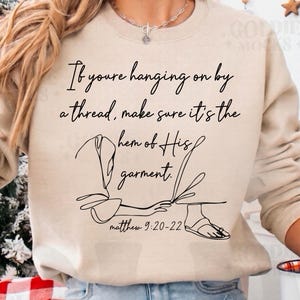 May include: Beige sweatshirt with the text "If you're hanging on by a thread, make sure it's the hem of His garment." A line drawing of a person's feet and garment is below the text. The Bible verse Matthew 9:20-22 is at the bottom.