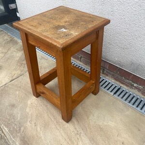 May include: A wooden stool with a square top and a simple design. The top is made of light brown wood with a darker brown stain. The stool has four legs and a simple frame.
