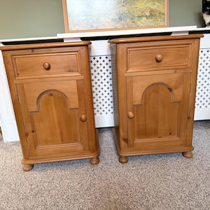 May include: A pair of wooden bedside cabinets. Each cabinet features a drawer with a round knob, a door with an arched panel, and a small knob. The cabinets are made of light-coloured wood and stand on small, rounded feet.