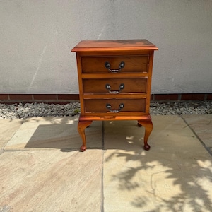 May include: A wooden bedside table with a warm brown finish, featuring three horizontal drawers stacked vertically. Each drawer is equipped with two dark metal swing handles. The piece stands on four curved cabriole legs.
