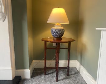 Puede incluir: Una mesa de madera con una tapa octogonal y una lámpara blanca con una base de patrón azul y blanco. La lámpara está sobre la mesa y la mesa está en una esquina de una habitación.