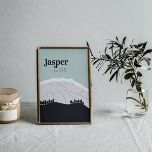 Peut inclure: Une impression encadrée dorée avec le mot "jasper" et un paysage de montagne stylisé dans des tons de bleu, rose et blanc. L'impression est posée sur une nappe en lin blanc avec un pot de bougie et un vase de verdure.