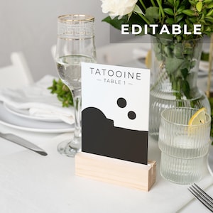 Puede incluir: Una tarjeta de número de mesa en blanco y negro con el texto "Tatooine - Mesa 1 -" y un gráfico de un círculo negro y una luna creciente negra. La tarjeta está en un soporte de madera.