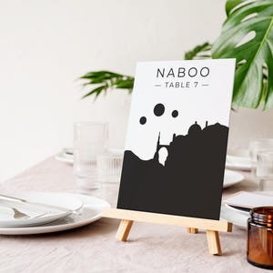 May include: A black and white table number card with the text "NABOO - TABLE 7" and a silhouette of a city with three moons. The card is on a small wooden easel.