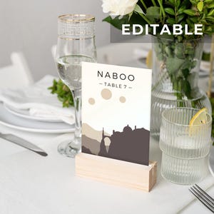 以下が含まれることがあります： 「NABOO - TABLE 7 -」というテキストと、3 つの月のある都市のシルエットが描かれた白いテーブル番号カード。カードは木製スタンドの上に置かれています。