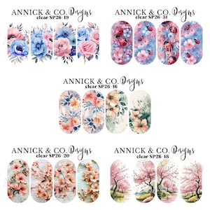 Peut inclure: Planches de décalcomanies pour ongles avec des motifs floraux et de fleurs de cerisier. Les décalcomanies présentent des fleurs à l'aquarelle dans les tons de rose, de bleu et de pêche, ainsi que des coccinelles. La marque "ANNICK & CO. Designs" est visible.