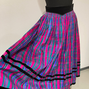 Op de afbeelding: Een vintage geruite rok in roze, turquoise en blauw met verticale strepen. Het heeft zwarte fluwelen afwerking aan de taille en zoom. De rok wordt op een mannequin gepresenteerd. Het ontwerp doet denken aan de jaren 80.