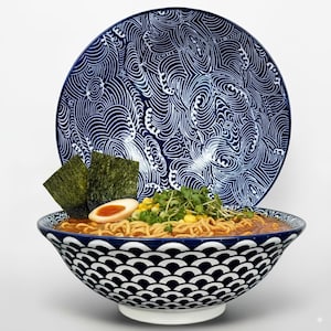 Pode incluir: Uma tigela de ramen com macarrão, caldo, algas marinhas e um ovo, apresentada em uma tigela com estampa azul e branca. Um prato combinando com um desenho de ondas está no fundo. A tigela fica em uma base branca.
