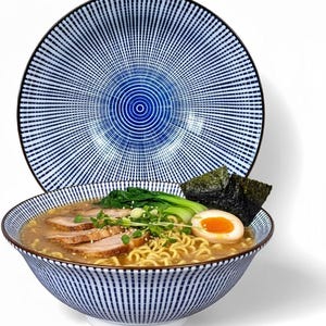 Può includere: Una ciotola di ramen con noodles, fette di maiale, un uovo alla coque e alghe. La ciotola ha un motivo a righe blu e bianche, e un piatto abbinato è dietro. Il cibo è disposto su una superficie bianca.