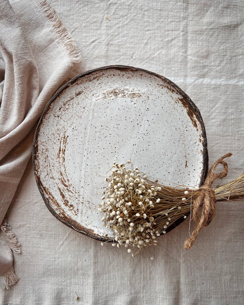 Pu&ograve; includere: Un piatto in ceramica bianca con macchie marroni e una finitura rustica. Un mazzo di fiori bianchi secchi con steli marroni riposa sul piatto.