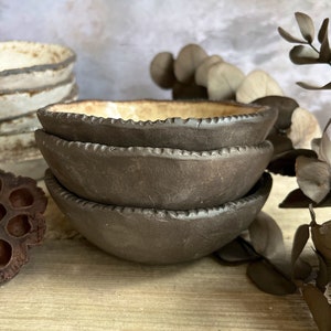 May include: Three stacked brown ceramic bowls with a textured rim. The bowls are on a wooden surface with a light brown background.