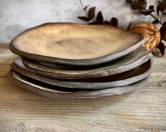 May include: A stack of four handmade ceramic plates. The plates have a unique, organic shape with a dark brown rim and a light brown center. The plates are stacked on top of each other, showcasing their individual forms and textures. The plates are on a wooden surface.
