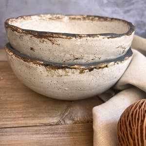 May include: Two white ceramic bowls with a speckled glaze and a dark brown rim. The bowls are stacked on top of each other and are sitting on a wooden surface.