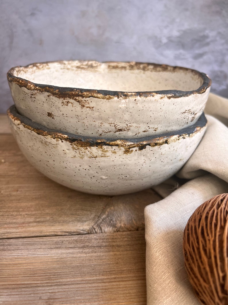 May include: Two white ceramic bowls with a speckled glaze and a dark brown rim. The bowls are stacked on top of each other and are sitting on a wooden surface.