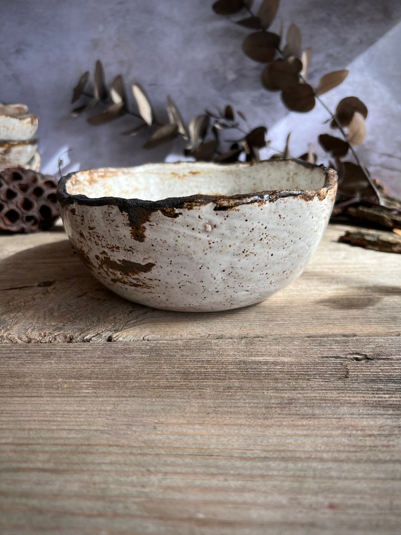 May include: A handmade ceramic bowl with a speckled white glaze and a dark brown rim. The bowl has a rustic, organic shape and sits on a wooden surface. Dried foliage and other natural elements are in the background.