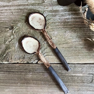 May include: Two ceramic spoons with a white glaze and black rim. The spoons are on a wooden surface.