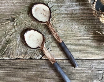Könnte beinhalten: Zwei Keramikschöpfer mit weißem Überzug und schwarzem Rand. Die Schöpfer liegen auf einer Holzoberfläche.