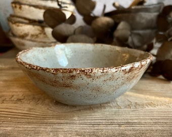 May include: A small, light blue ceramic bowl with a speckled brown glaze. The bowl has a slightly irregular rim and is sitting on a wooden surface. "Style 1" is written in yellow text on the surface.