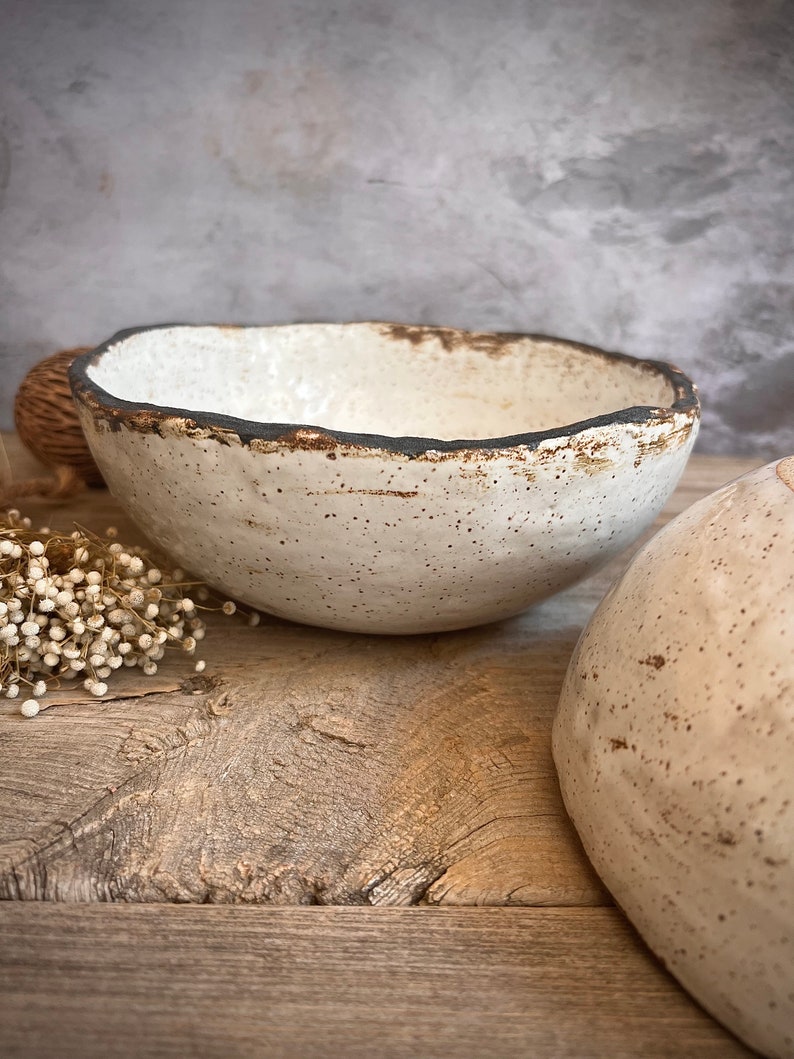 May include: A white ceramic bowl with a dark brown rim and speckled glaze. The bowl is sitting on a wooden surface.