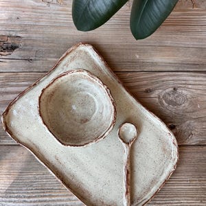 May include: A handmade ceramic serving set featuring a rectangular tray, a small bowl, and a spoon. The set is off-white with brown speckles and edges. The rustic design is perfect for serving appetizers or small dishes.