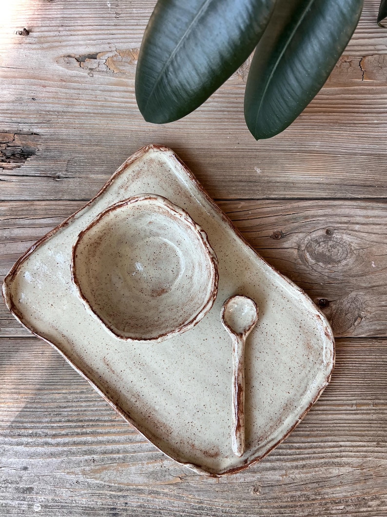 May include: A handmade ceramic serving set featuring a rectangular tray, a small bowl, and a spoon. The set is off-white with brown speckles and edges. The rustic design is perfect for serving appetizers or small dishes.