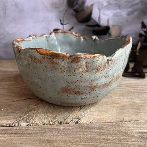 May include: A handmade ceramic bowl with a unique, organic shape. The bowl features a light blue-grey glaze with a textured surface and a contrasting brown rim. The rustic design adds a touch of natural elegance to any kitchen or dining space.
