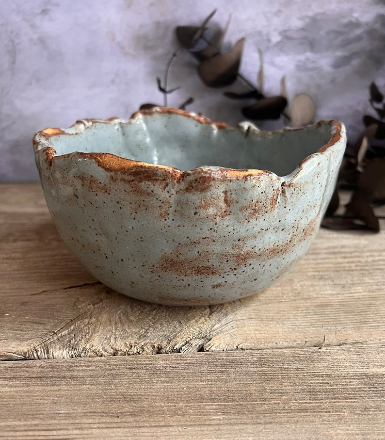 May include: A handmade ceramic bowl with a unique, organic shape. The bowl features a light blue-grey glaze with a textured surface and a contrasting brown rim. The rustic design adds a touch of natural elegance to any kitchen or dining space.