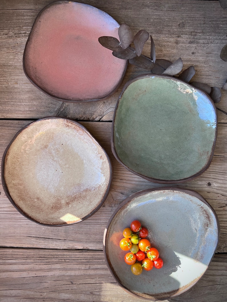 May include: Four ceramic plates in different colors and textures. The plates are arranged on a wooden surface. One plate has a small pile of red and yellow cherry tomatoes.
