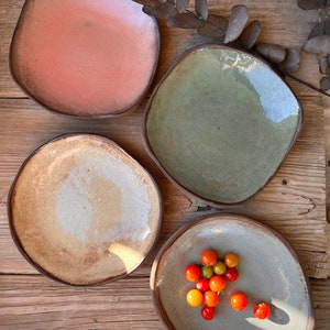 May include: Four ceramic plates in different colors and textures. The plates are arranged on a wooden surface. One plate has a light brown glaze, another has a light green glaze, another has a light pink glaze, and the last plate has a light gray glaze and is filled with small red and orange tomatoes.