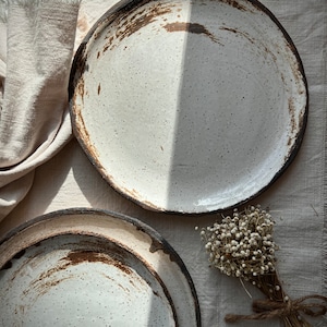Pu&ograve; includere: Due piatti in ceramica bianca con smalto macchiato di marrone e un bordo marrone scuro. I piatti sono impilati uno sopra l'altro. Un piccolo mazzo di fiori bianchi secchi &egrave; sullo sfondo.