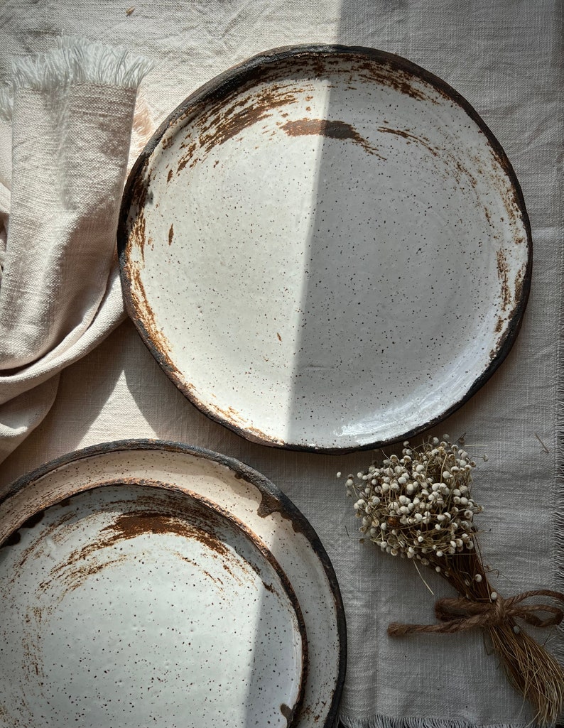 Pu&ograve; includere: Due piatti in ceramica bianca con smalto macchiato di marrone e un bordo marrone scuro. I piatti sono impilati uno sopra l'altro. Un piccolo mazzo di fiori bianchi secchi &egrave; sullo sfondo.