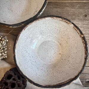 May include: Two white ceramic bowls with a speckled glaze and a dark brown rim. The bowls are sitting on a light brown wooden surface.