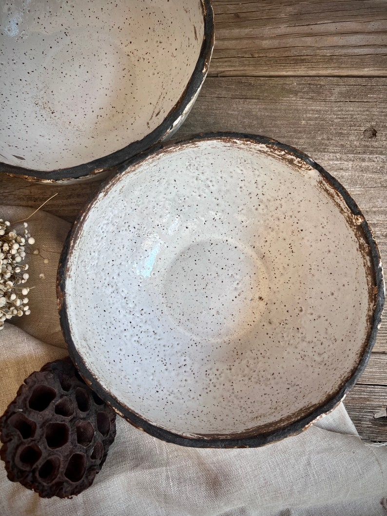May include: Two white ceramic bowls with a speckled glaze and a dark brown rim. The bowls are sitting on a light brown wooden surface.