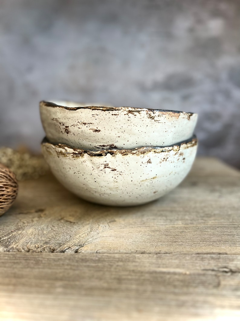 May include: Two white ceramic bowls with a speckled finish and a brown rim. The bowls are stacked on top of each other and are sitting on a wooden surface.