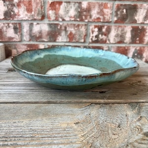 May include: A shallow, round ceramic bowl with an irregular, wavy rim. The interior features a gradient of colors, transitioning from a creamy white center to shades of teal, blue, and brown along the edges, with a glossy, reactive glaze finish.
