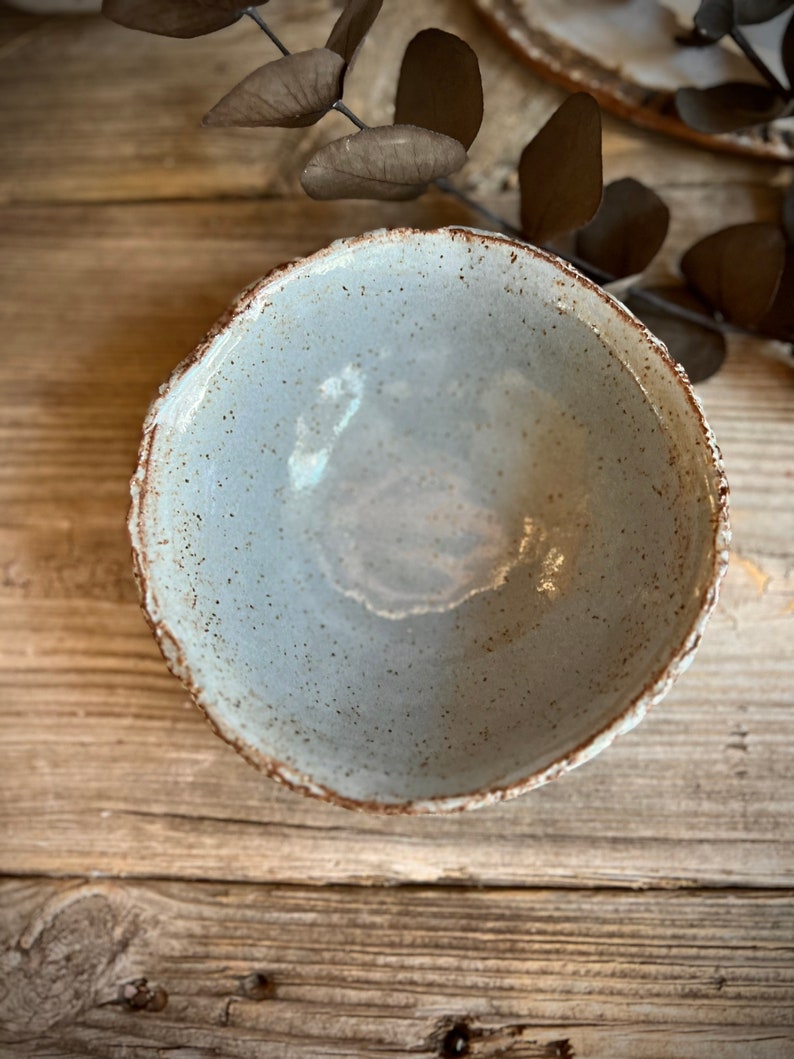May include: A light gray ceramic bowl with a speckled glaze and a slightly uneven rim. The bowl is empty and sitting on a wooden surface.