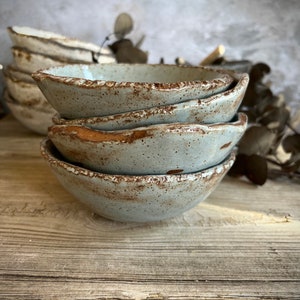 May include: A stack of four small, rustic, light blue ceramic bowls with a speckled glaze. The bowls have a slightly uneven, handmade appearance and are stacked on a weathered wooden surface.