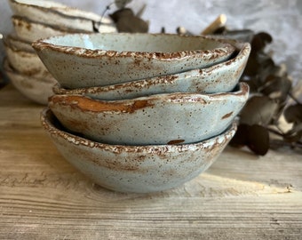 May include: A stack of four small, rustic, light blue ceramic bowls with a speckled glaze. The bowls have a slightly uneven, handmade appearance and are stacked on a weathered wooden surface.