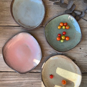 May include: Four ceramic plates in different colors and shapes. The plates are on a wooden surface. One plate has a green glaze and is filled with small red and orange tomatoes.