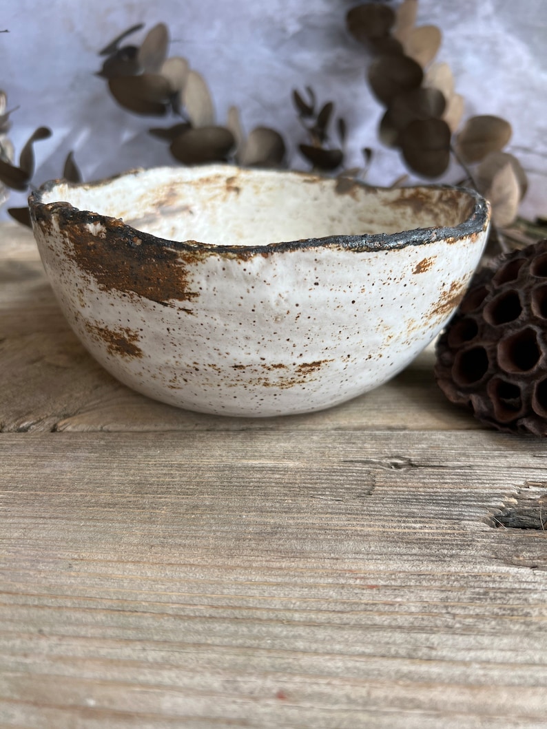 May include: A handmade ceramic bowl with a textured white interior and a dark brown, rough-edged rim. The bowl has a rustic, speckled appearance and sits on a wooden surface. Dried foliage and a seed pod are in the background.