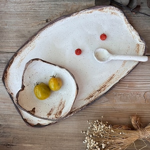 May include: A set of handmade ceramic serving dishes. The set includes a large rectangular platter, a smaller irregular-shaped bowl, and a small spoon. The dishes are white with brown edges and speckled details. Two small yellow tomatoes and red berries are on the dishes.