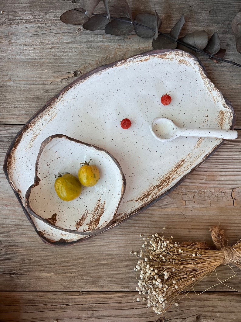 May include: A set of handmade ceramic serving dishes. The set includes a large rectangular platter, a smaller irregular-shaped bowl, and a small spoon. The dishes are white with brown edges and speckled details. Two small yellow tomatoes and red berries are on the dishes.