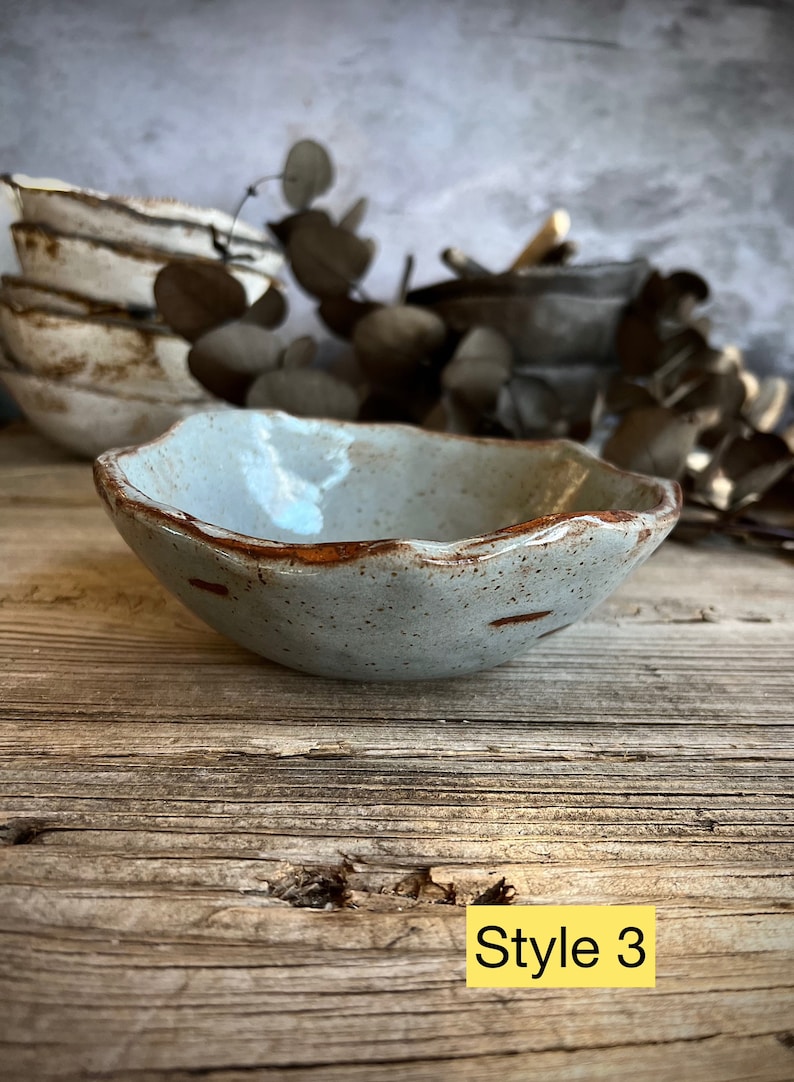 May include: A small, light blue ceramic bowl with a brown rim. The bowl has a speckled glaze and a slightly irregular shape. The bowl is sitting on a wooden surface. Style 3.