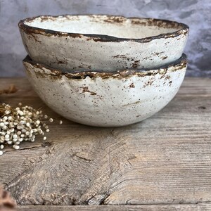 May include: Two white ceramic bowls with a rustic, speckled glaze and a brown rim. The bowls are stacked on top of each other on a wooden surface.