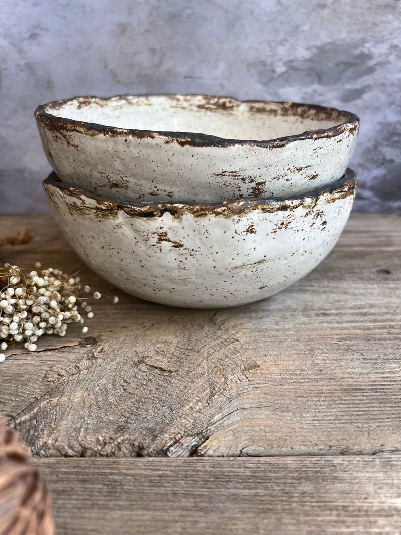May include: Two white ceramic bowls with a rustic, speckled glaze and a brown rim. The bowls are stacked on top of each other on a wooden surface.