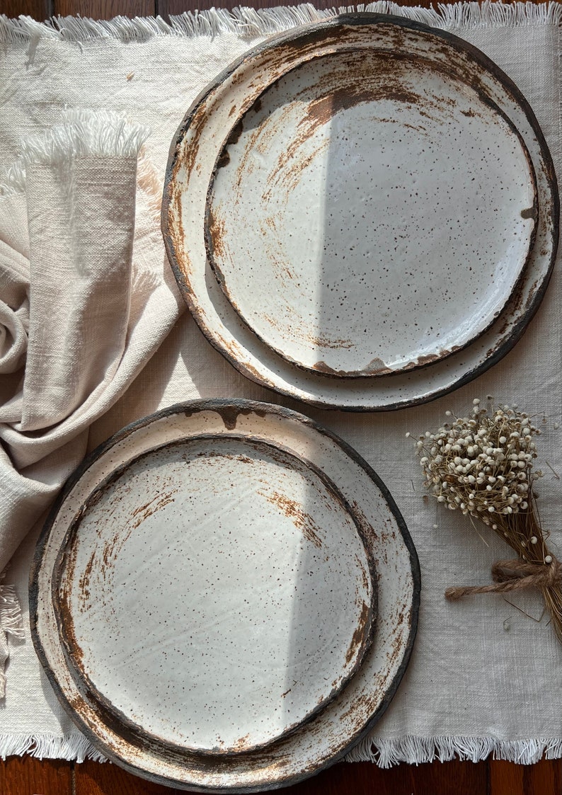 Pu&ograve; includere: Due piatti in ceramica bianca con una glassa macchiettata e un bordo marrone. I piatti sono annidati e hanno un aspetto rustico e fatto a mano.