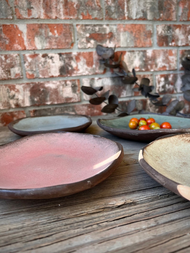 May include: Four ceramic plates in different colors and shapes, including a light blue, a light pink, a light green, and a beige plate. The plates are arranged on a wooden surface.