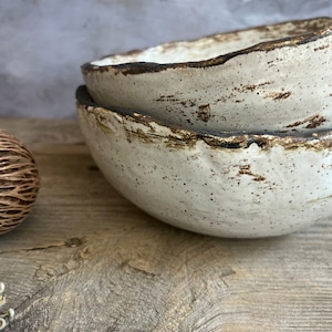 May include: Two white ceramic bowls with a speckled brown glaze and a dark brown rim. The bowls are stacked on top of each other and are sitting on a wooden surface.