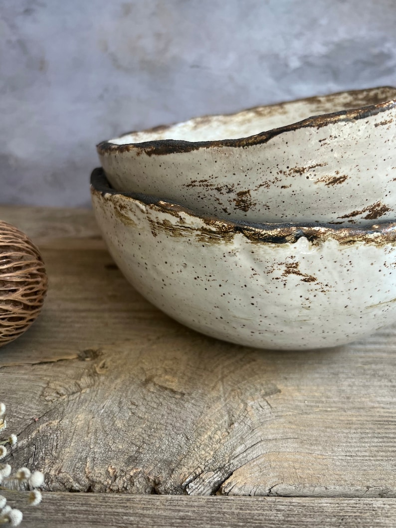 May include: Two white ceramic bowls with a speckled brown glaze and a dark brown rim. The bowls are stacked on top of each other and are sitting on a wooden surface.